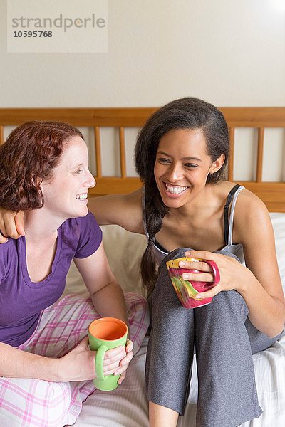 Junge Frauen sitzen auf dem Bett  Arm um Freund  halten Tasse lächelnd