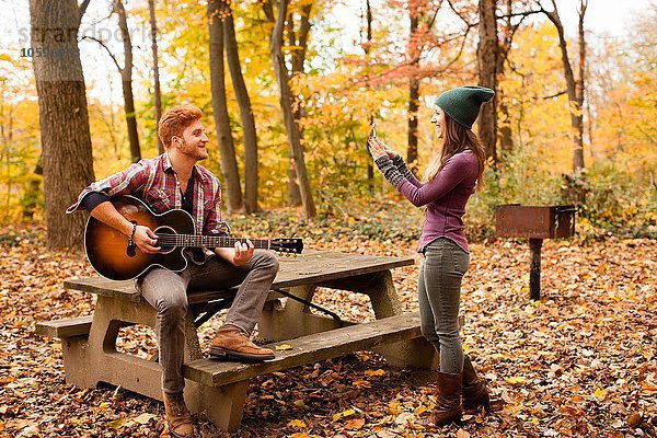 Junge Frau fotografiert Gitarre spielenden Freund im Herbstwald