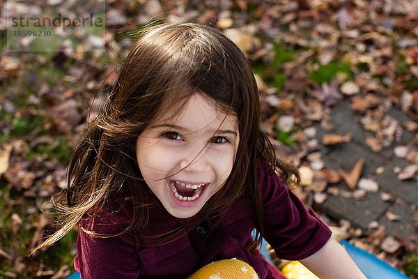 Hochwinkliges Porträt eines jungen Mädchens zwischen Herbstblättern mit offenem Mund und lächelndem Blick auf die Kamera.