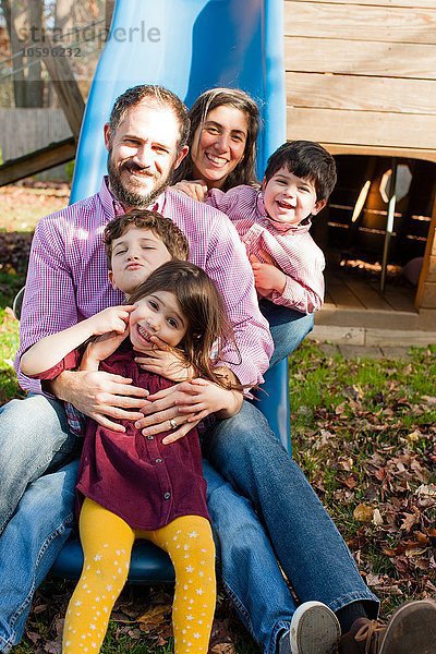 Familie in einer Reihe zwischen den Beinen auf einer Spielplatzrutsche  mit lächelndem Blick auf die Kamera.