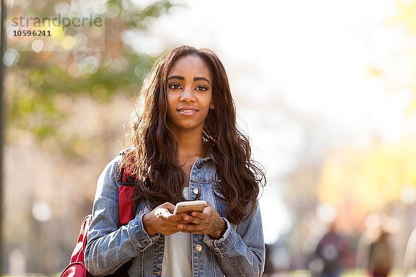 Junge Frau mit langen braunen Haaren  die eine Jeansjacke trägt  die ein Smartphone hält und lächelnd wegsieht.