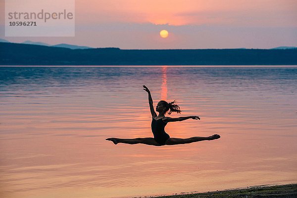 Seitenansicht des Mädchens am Meer bei Sonnenuntergang  das in der Mitte der Luft springt  die Arme erhoben  um die Spaltungen zu machen