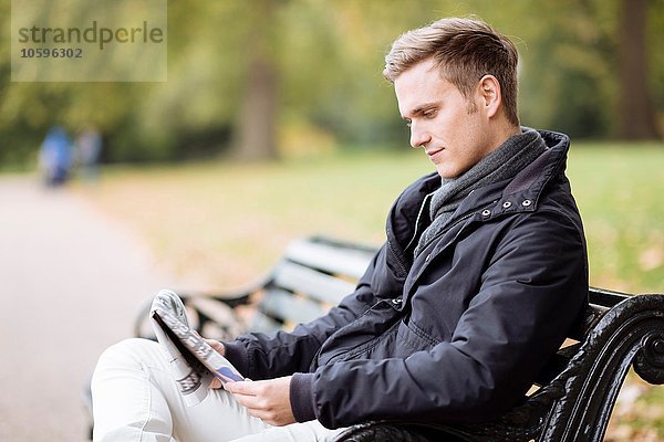Junger Mann sitzt auf der Bank und liest Zeitung im Park.