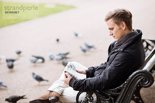 Junger Mann sitzt auf der Parkbank und liest Zeitung.