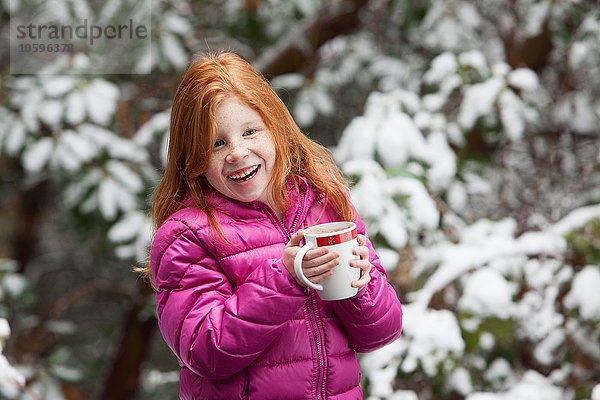 Rothaariges Mädchen vor schneebedeckten Bäumen  mit rosa gepolstertem Mantel  der den Becher hält und lächelnd wegsieht.