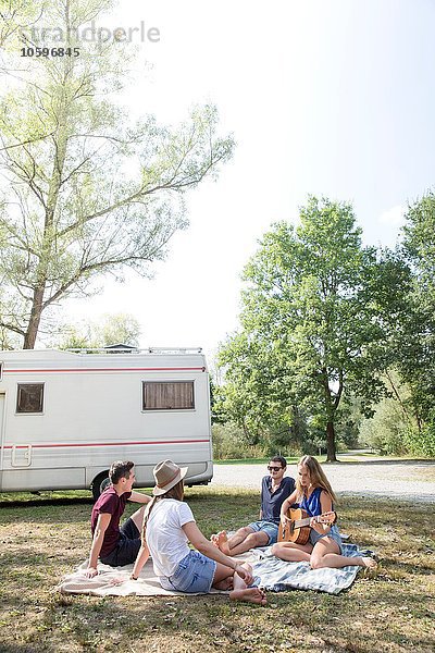 Gruppe junger Erwachsener auf Picknickdecke sitzend  entspannend  Wohnmobil im Hintergrund