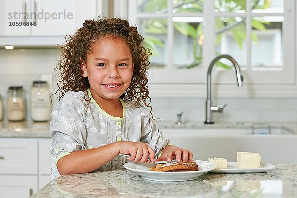 Mädchen an der Küchentheke streicht Butter auf Toast und sieht lächelnd in die Kamera.