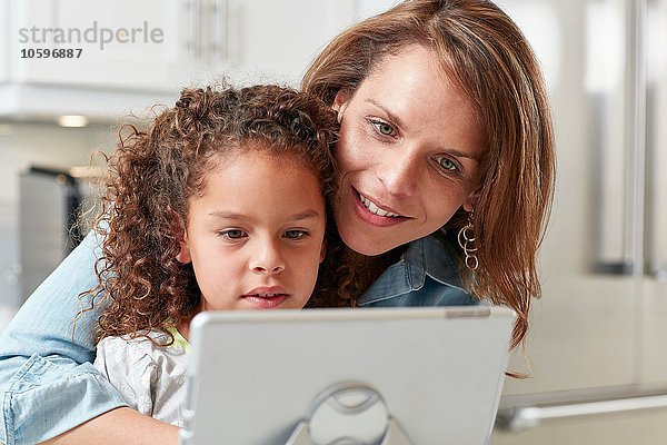 Mutter mit Arm um Tochter mit digitalem Tablett und lächelndem Blick nach unten