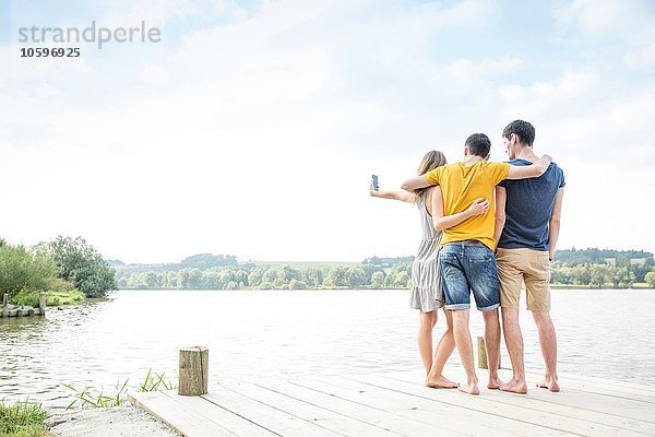 Drei junge Erwachsene am Steg stehend  Selbstporträt  mit Smartphone  Rückansicht
