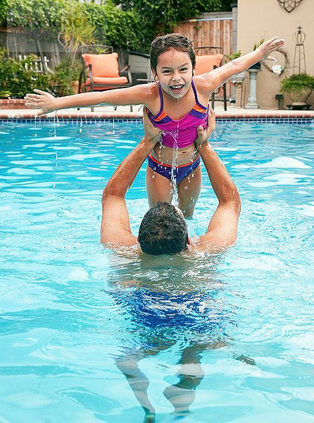 Vater im Schwimmbad hebt Tochter hoch  Arme geöffnet  Kamera lächelnd betrachtend