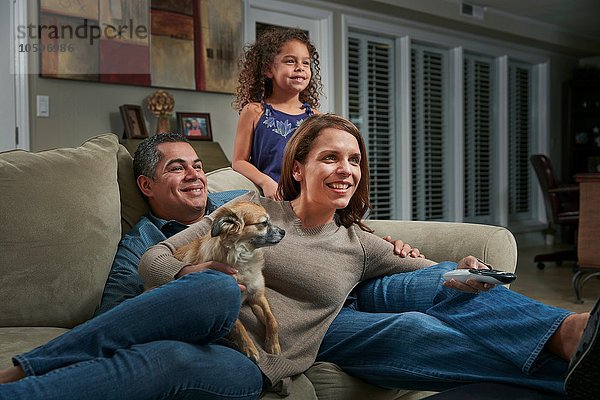 Eltern und Mädchen im Wohnzimmer auf dem Sofa mit einem Hund  der lächelnd wegguckt.