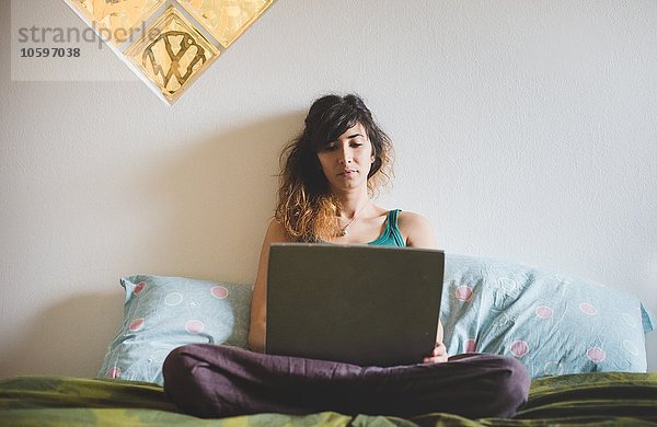 Vorderansicht der mittleren erwachsenen Frau  die mit einem Laptop im Kreuzbein auf dem Bett sitzt.