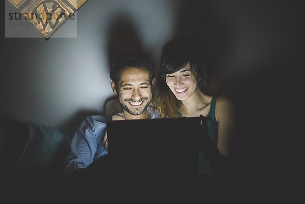 Vorderansicht des jungen Paares  beleuchtet von einem Laptop-Bildschirm  mit lächelndem Blick nach unten.