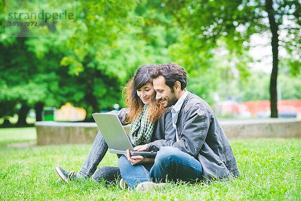 Junges Paar sitzt im Schneidersitz auf Gras mit Laptop und sieht lächelnd nach unten.