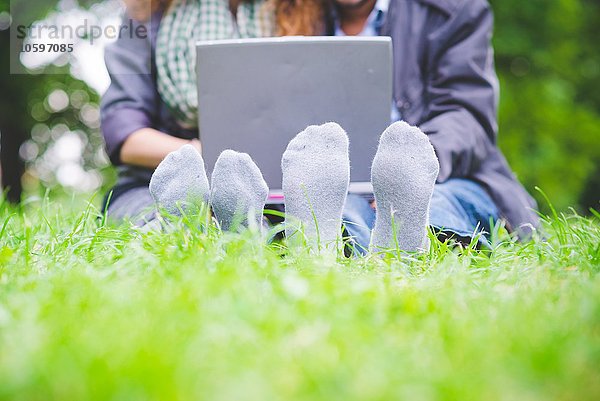 Füße eines jungen Paares mit Socken  die auf Gras sitzen und einen Laptop benutzen