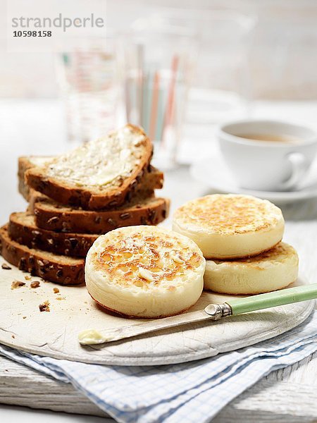 Crumpets und entkerntes Vollkornbrot mit Butter auf Vintage-Schneidebrett mit Tasse Tee