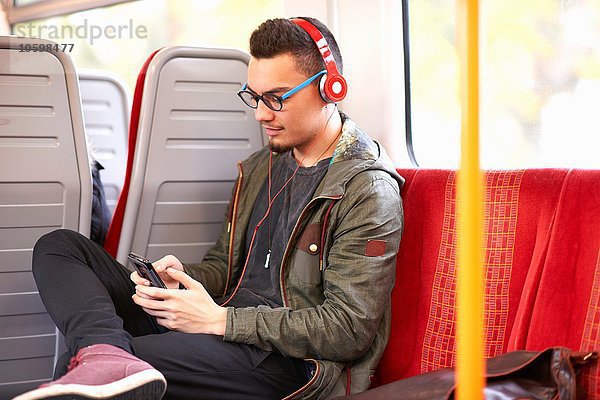 Junger Mann sitzt im Zug  benutzt Smartphone  trägt Kopfhörer