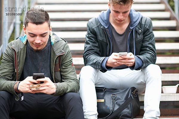 Zwei junge Männer  auf Stufen sitzend  mit Smartphones  im Freien