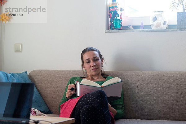 Vorderansicht der mittleren erwachsenen Frau  die auf dem Sofa sitzt und auf das Lesebuch schaut.