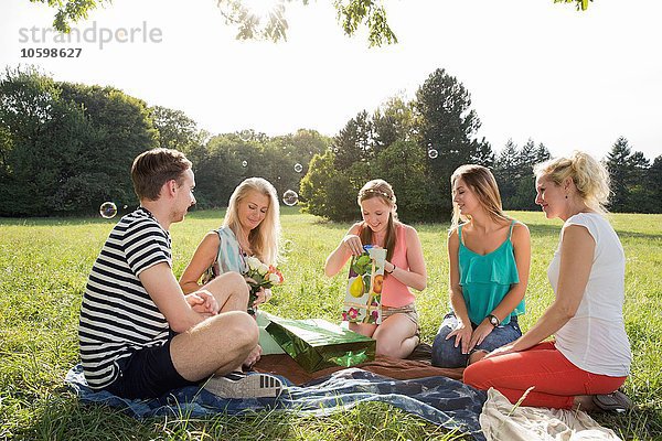 Familie sitzt auf Picknickdecke im Park und schenkt reife Frau Blumen und Geschenke.