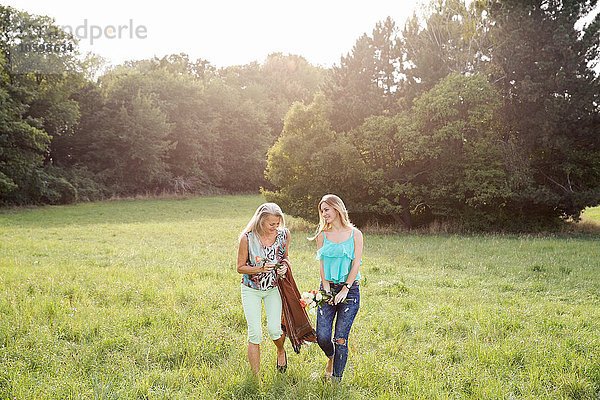 Vorderansicht von Frauen mit Picknickdecke und Blumen  die lächelnd über die Feldlandschaft gehen.