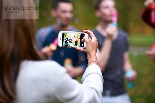 Über Schulteransicht einer jungen Frau  die ein Smartphone-Foto von Freunden im Park macht.