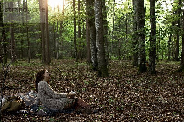 Seitenansicht einer reifen Frau  die im Wald auf einem Picknickteppich sitzt und eine Kaffeetasse mit Blick in die Ferne hält.