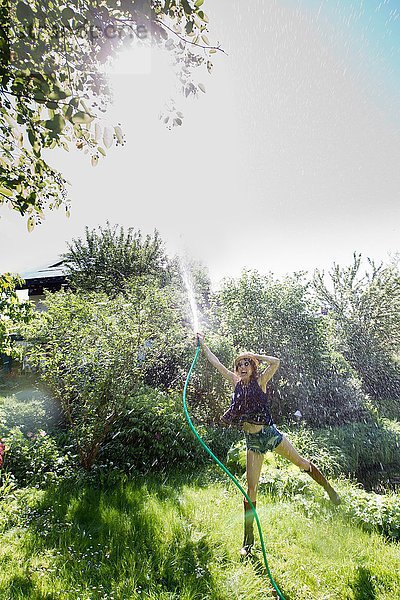 Vorderansicht einer reifen Frau im Garten  die auf einem Bein steht und mit einem Schlauch Wasser in die Luft spritzt.