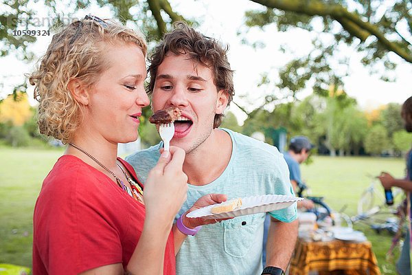 Fröhlicher junger Mann und Frau beim gemeinsamen Essen bei Sonnenuntergang im Park