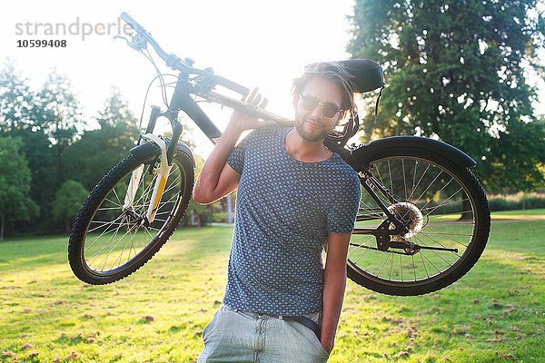 Porträt eines jungen Mannes  der bei Sonnenuntergang sein Fahrrad im Park trägt.