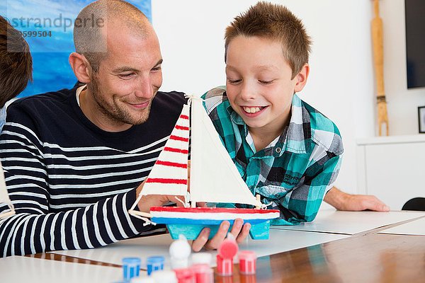 Mittlerer Erwachsener Mann und Sohn bewundern gemaltes Spielzeugboot am Küchentisch