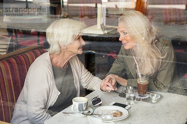 Mutter und Tochter sitzen zusammen im Café  halten sich an den Händen und sehen durch das Caféfenster.