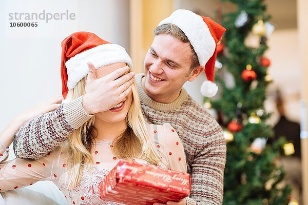 Junger Mann bedeckt die Augen der Freundin und überreicht ihr ein Weihnachtsgeschenk.