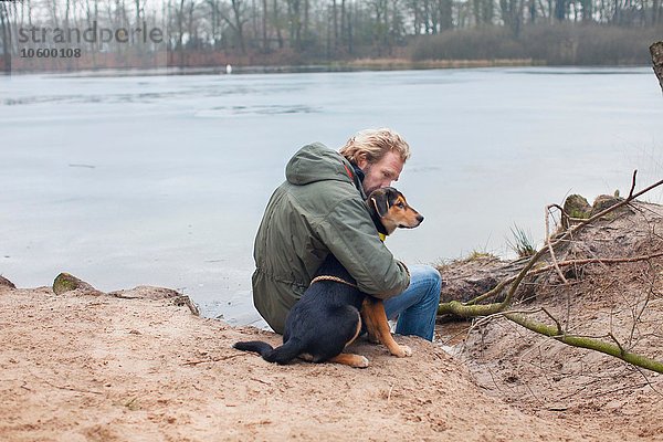 Erwachsener Mann umarmt Hund am Flussufer