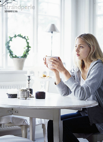 Junge Frau sitzt am Tisch und hält eine Tasse Kaffee