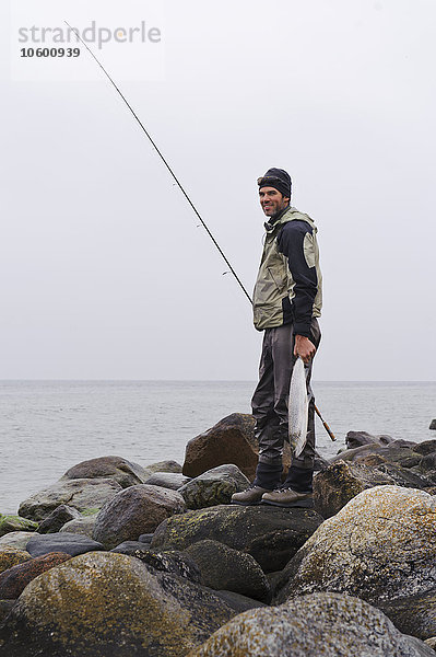 Lächelnder Mann steht an der Küste und hält Angelrute und Fisch