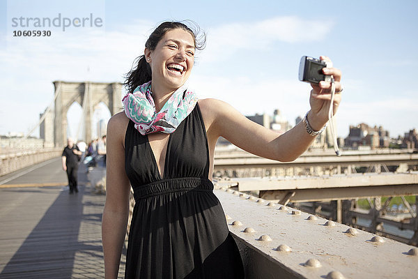 Frau im schwarzen Kleid macht Selfie mit Brooklyn Bridge im Hintergrund