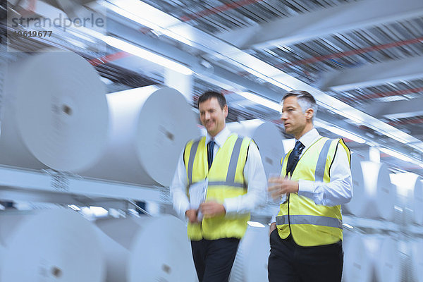 Geschäftsleute in reflektierender Kleidung  die in der Druckerei an großen Papierrollen entlanggehen