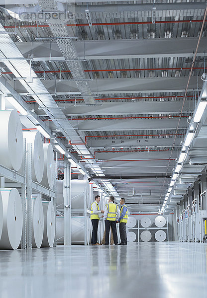 Arbeiter in reflektierender Kleidung reden in der Nähe von großen Papierrollen in der Druckerei