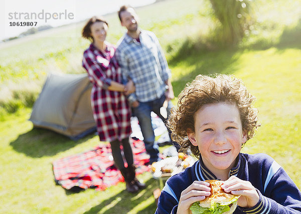 Portrait lächelnder Junge beim Hamburgeressen mit Eltern auf dem sonnigen Campingplatz