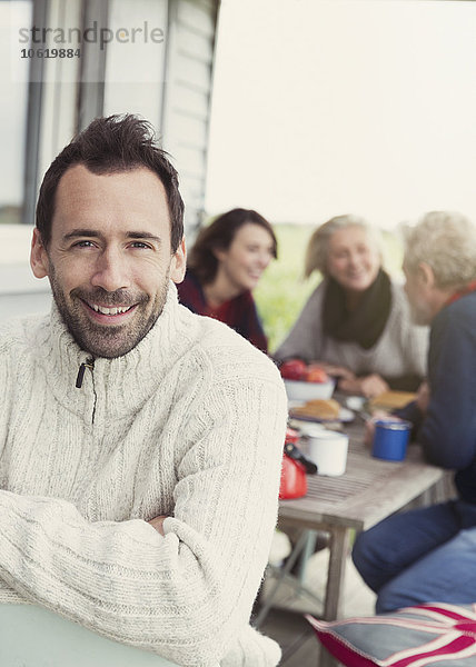Portrait lächelnder brünetter Mann im Pullover auf Veranda mit Familie im Hintergrund