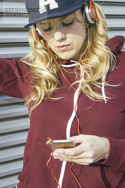 Blonde Frau mit Mütze und Kapuzenjacke beim Musikhören mit ihrem Smartphone