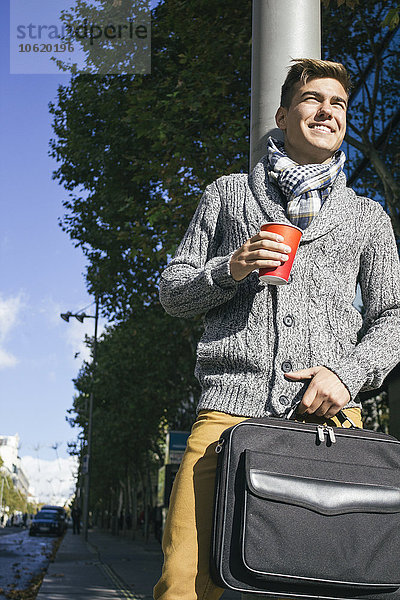 Junger Mann mit einem Aktenkoffer lächelnd  der einen Kaffee in einer Straße trinkt.
