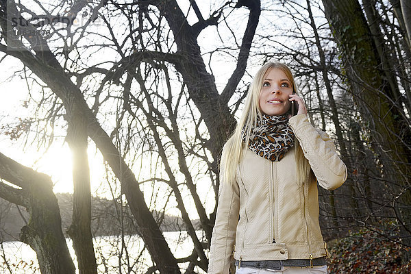 Porträt einer lächelnden blonden Frau beim Telefonieren mit dem Smartphone in der Natur
