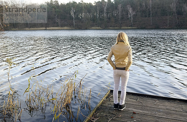 Rückansicht einer blonden Frau  die auf einem Holzsteg steht und einen See betrachtet.