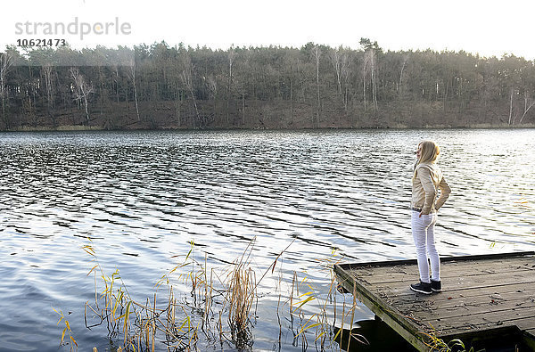 Blonde Frau steht auf einem Holzsteg und schaut auf einen See.
