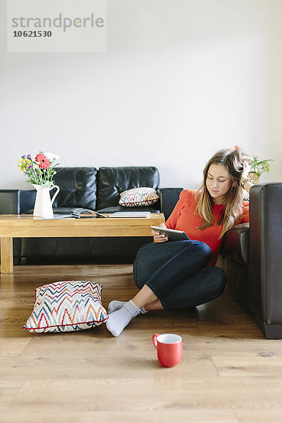 Junge Frau sitzt auf dem Boden ihres Wohnzimmers und schaut auf das digitale Tablett.