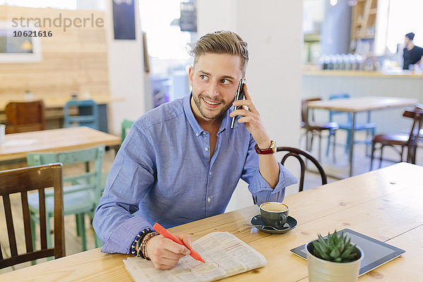 Mann mit Zeitung sitzt in einem Café und telefoniert mit dem Smartphone.