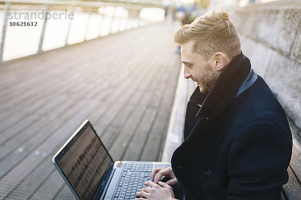 Junger Mann sitzt auf einer Bank und arbeitet mit einem Laptop.