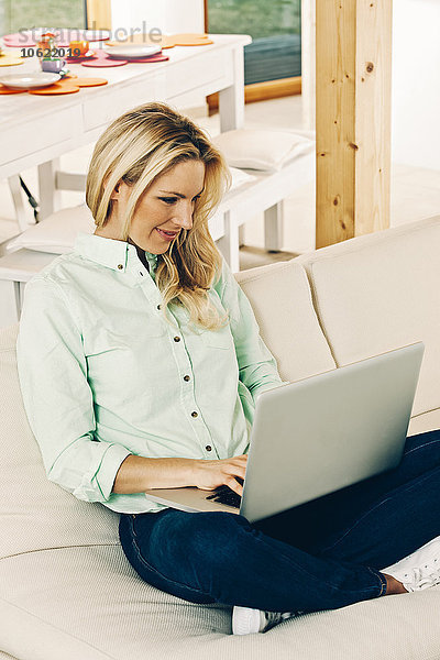 Lächelnde blonde Frau sitzt auf dem Sofa mit Laptop